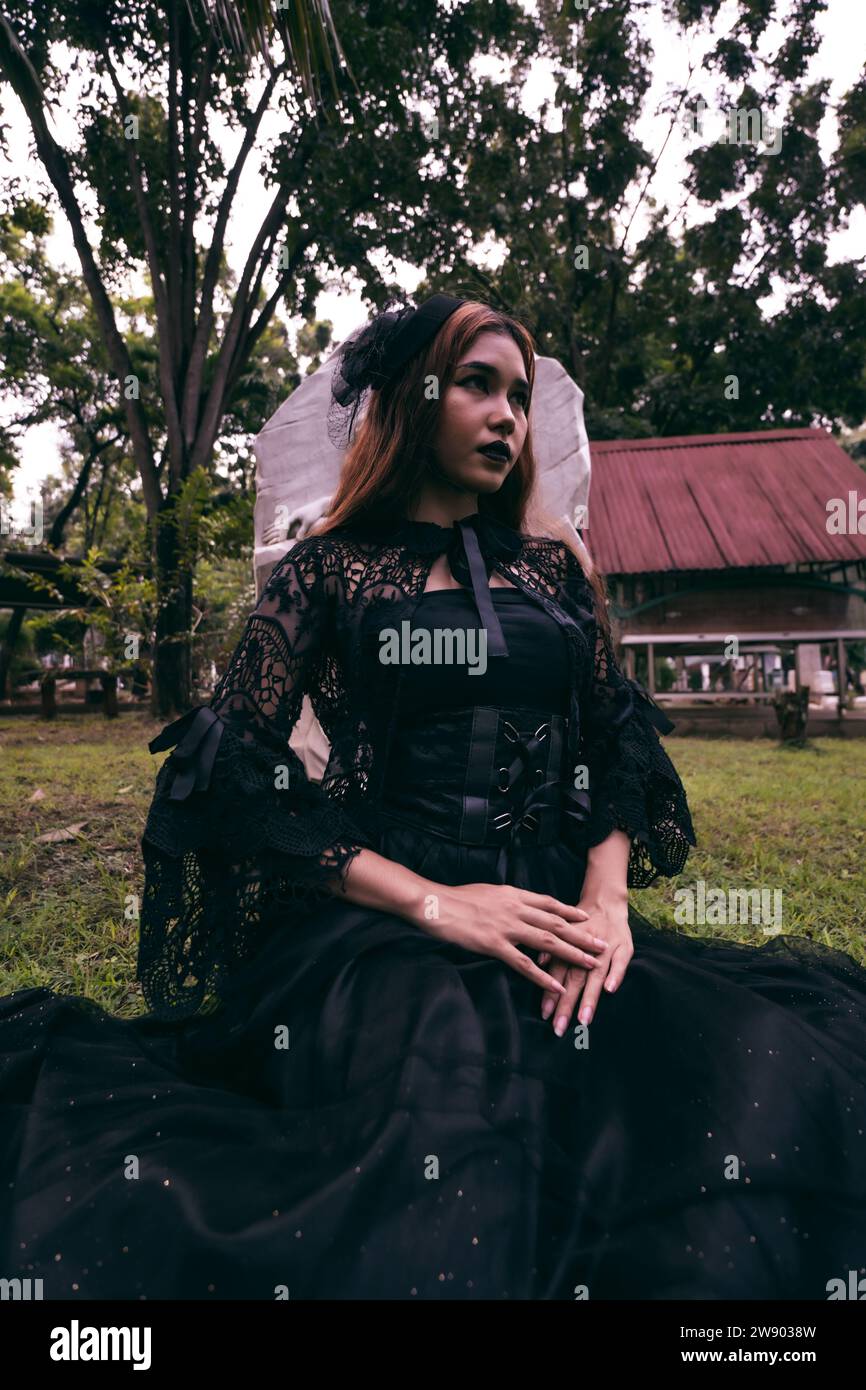a young witch in a long black dress and scary makeup was sitting in front of the tombstone in the afternoon Stock Photo