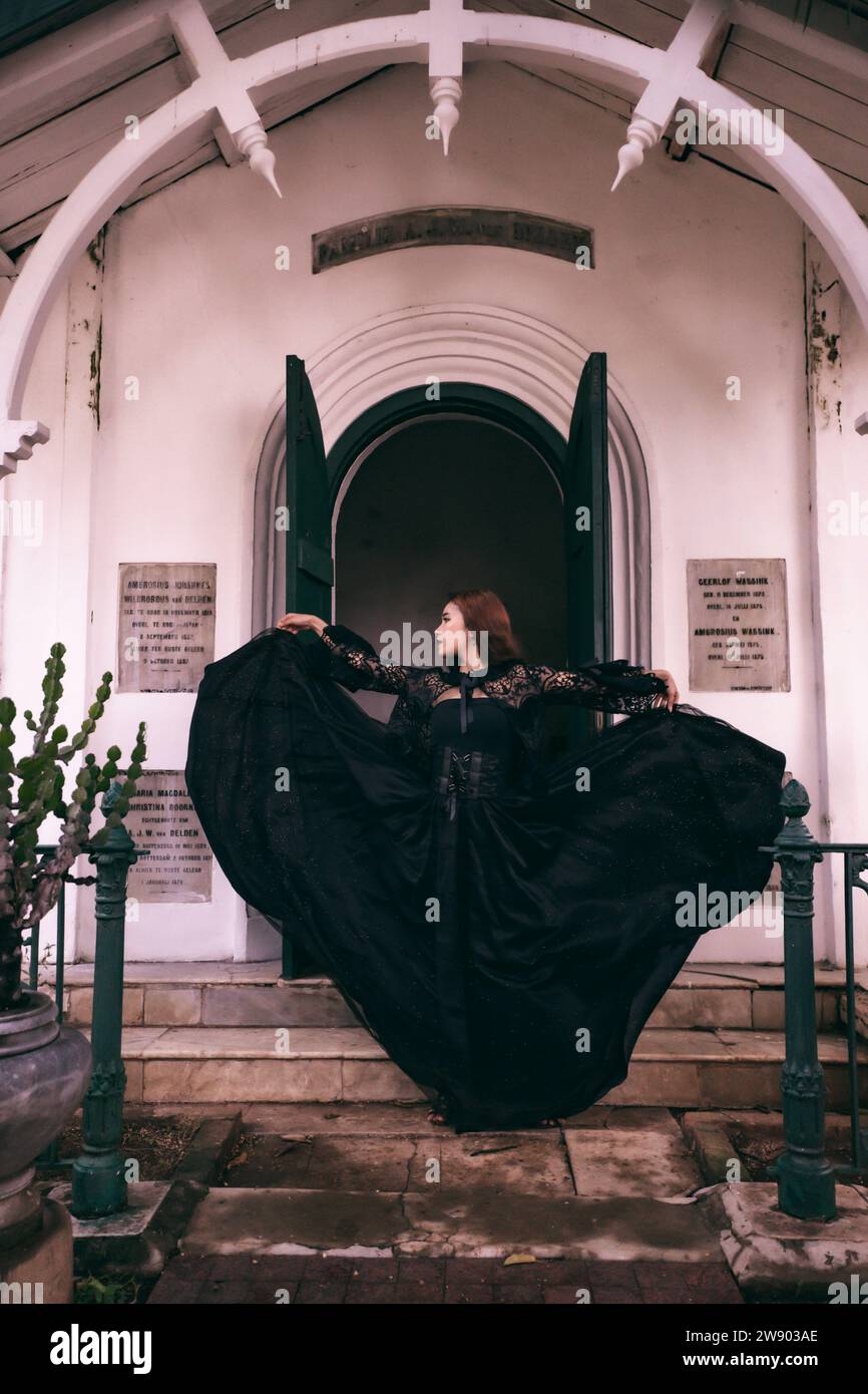 An elegant witch flicks her all black dress in the air in front of the cemetery gates after the Halloween festival begins in the afternoon Stock Photo