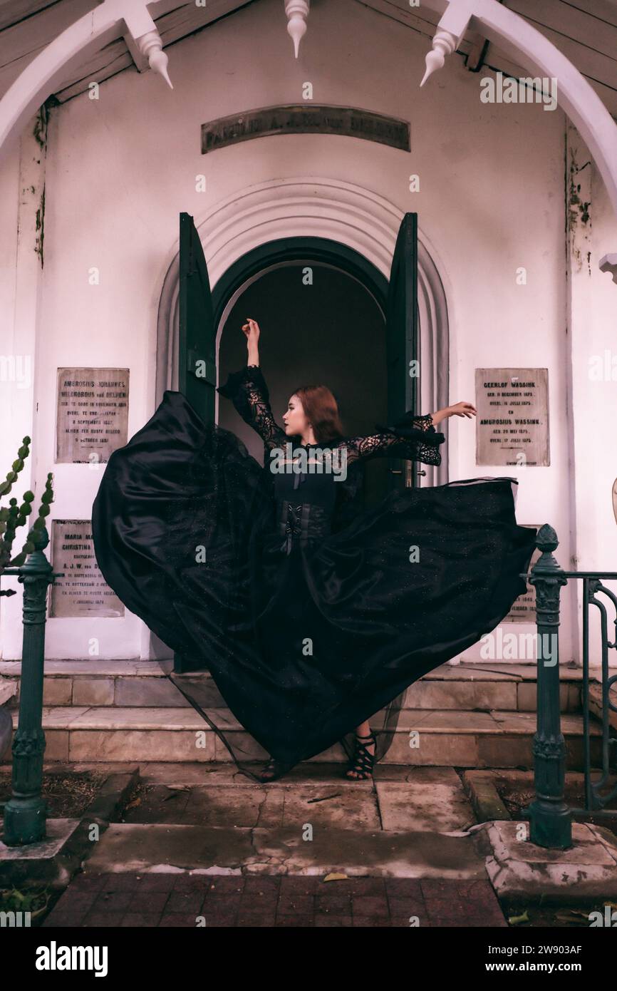 An elegant witch flicks her all black dress in the air in front of the cemetery gates after the Halloween festival begins in the afternoon Stock Photo