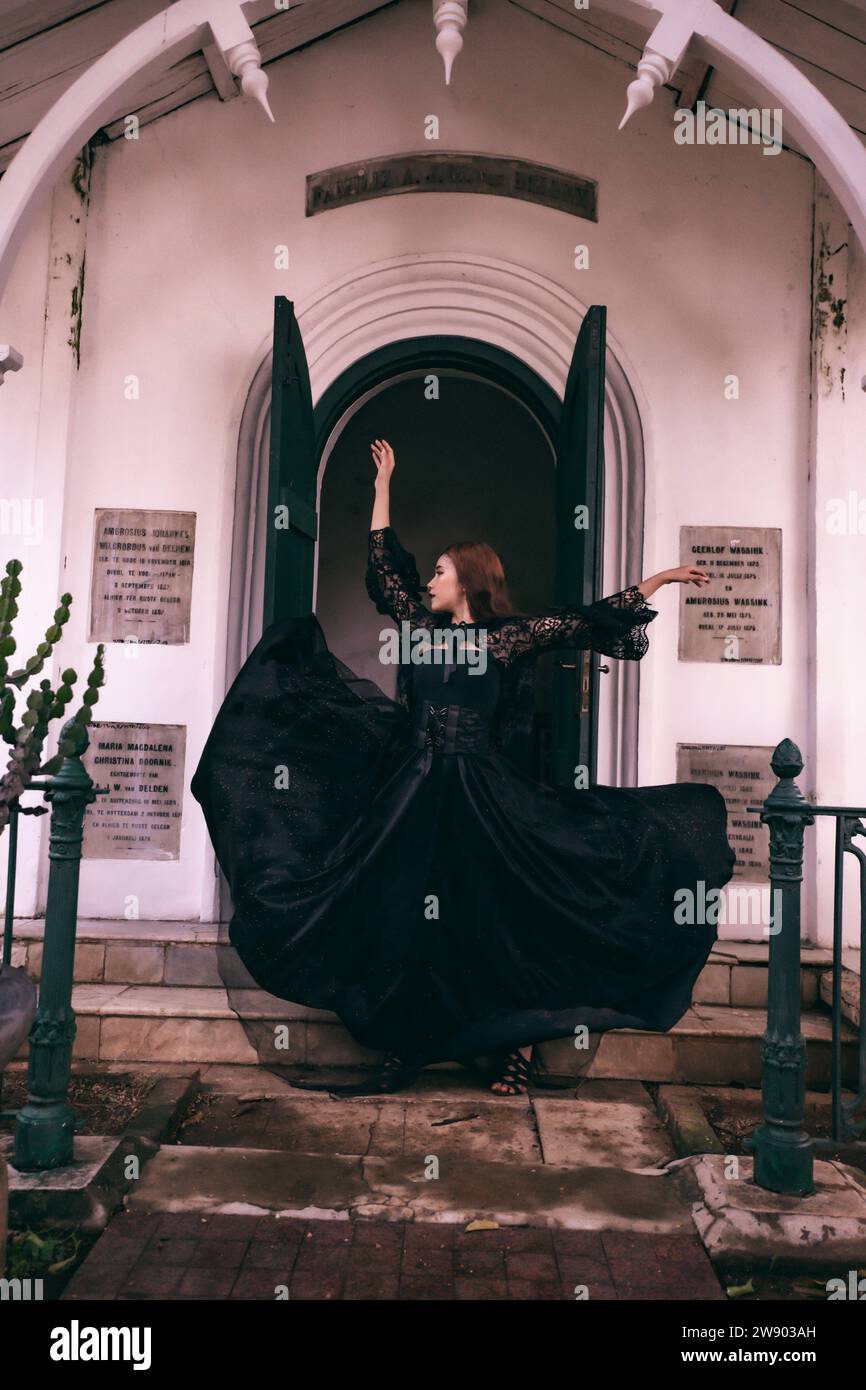 An elegant witch flicks her all black dress in the air in front of the cemetery gates after the Halloween festival begins in the afternoon Stock Photo