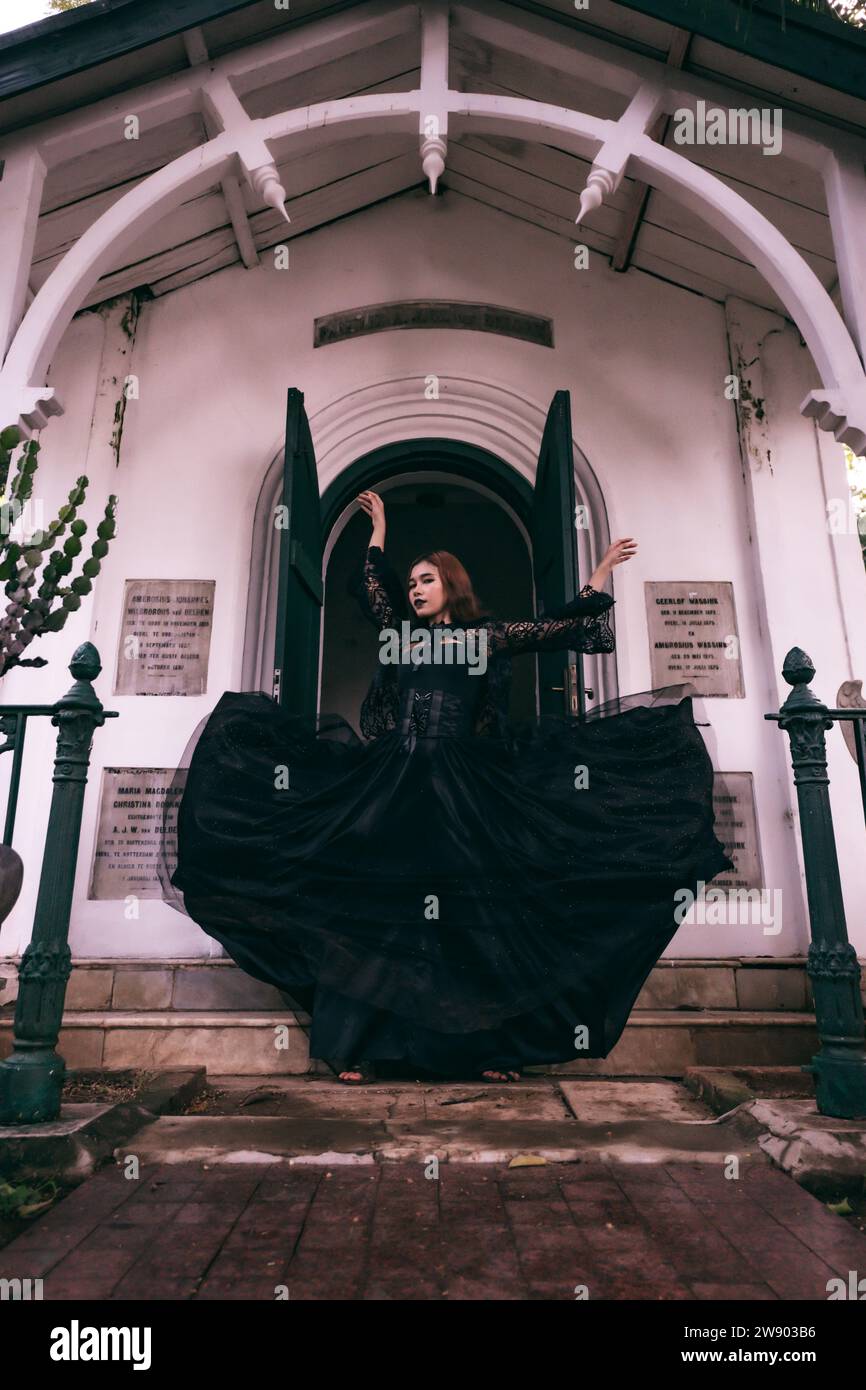 An elegant witch flicks her all black dress in the air in front of the cemetery gates after the Halloween festival begins in the afternoon Stock Photo