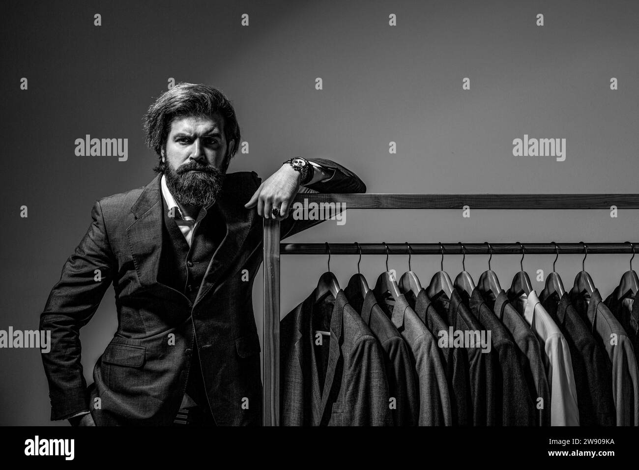 Handsome bearded fashion man in classical costume suit. Male suits hanging in a row. Black and white Stock Photo