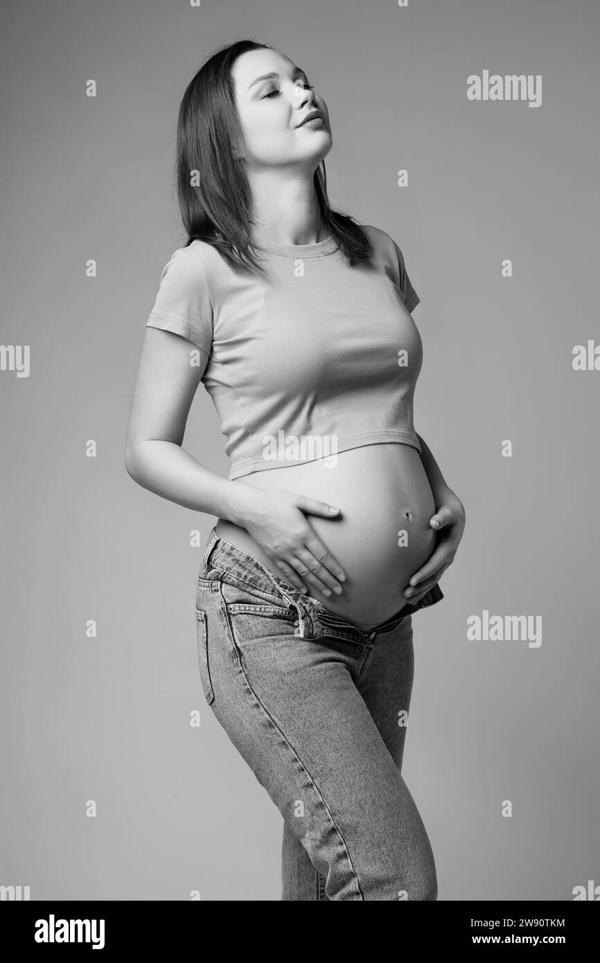 Black and white portrait of young pretty pregnant woman in t-shirt and jeans on gray background. Female belly exposed. 6th month of pregnancy. Eyes cl Stock Photo
