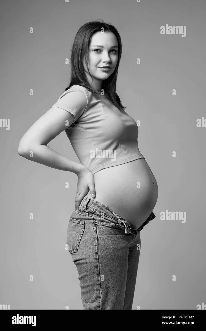 Black and white portrait of pretty pregnant woman in t-shirt and jeans on gray background. Female belly exposed. 6th month of pregnancy. Stock Photo