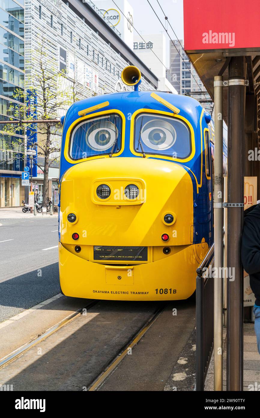 A tram, in the blue and yellow colours of Thomas Tank engine stopped at Okayama station, (Okayama Ekimae), tram stop. Daytime, sunny. Stock Photo