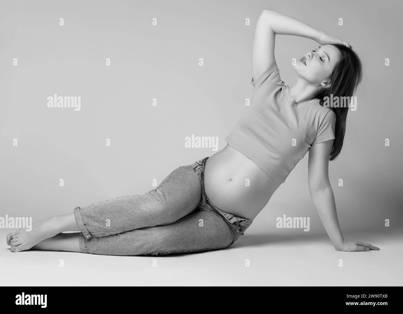 Black and white photo of young pretty pregnant woman in t-shirt and jeans sit on the floor on gray background. Female with belly exposed. 6th month of Stock Photo
