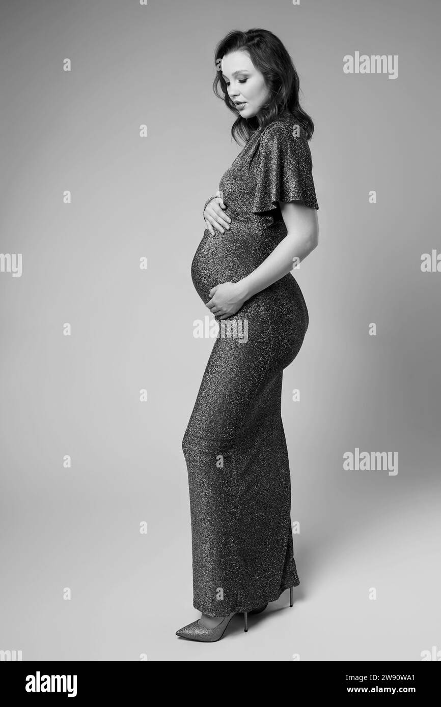 Black and white portrait of young pretty pregnant woman on gray studio background. Female in grey sequin dress with hands near pregnant belly. Stock Photo