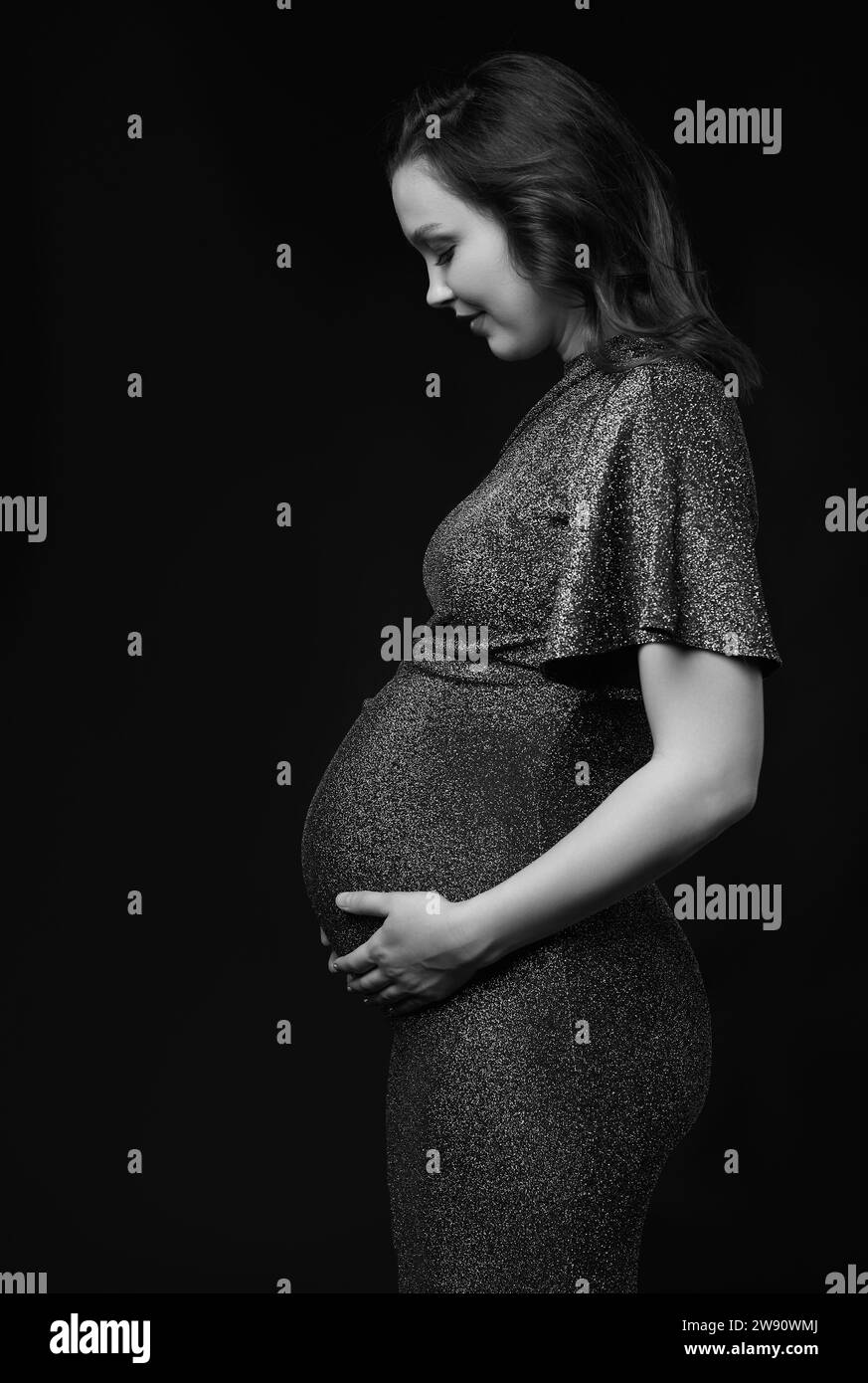 Black and white portrait of young pretty pregnant woman on black studio background. Female in grey sequin dress with hands near pregnant belly. Stock Photo