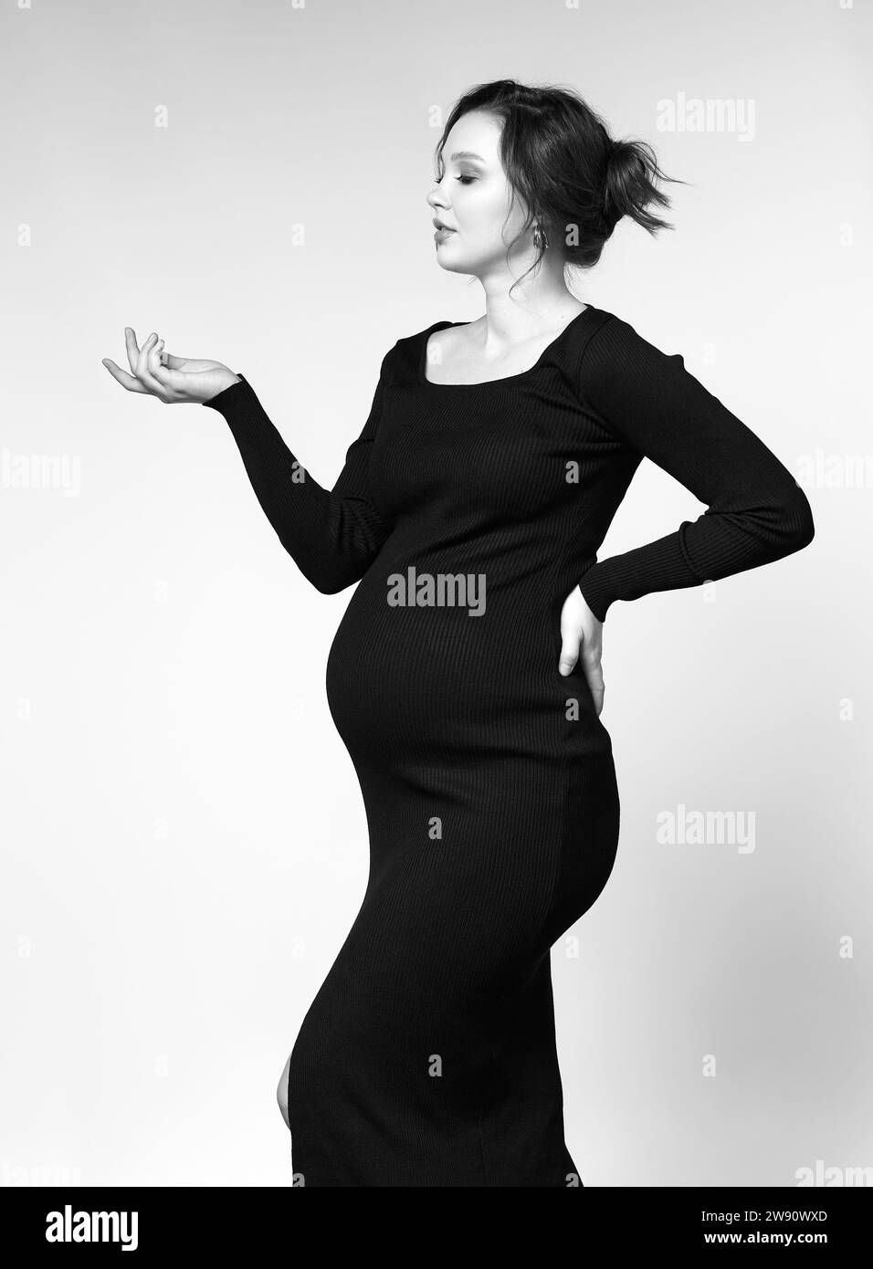 Black and white portrait of young pretty pregnant woman on gray studio background. Female in black dress. Stock Photo