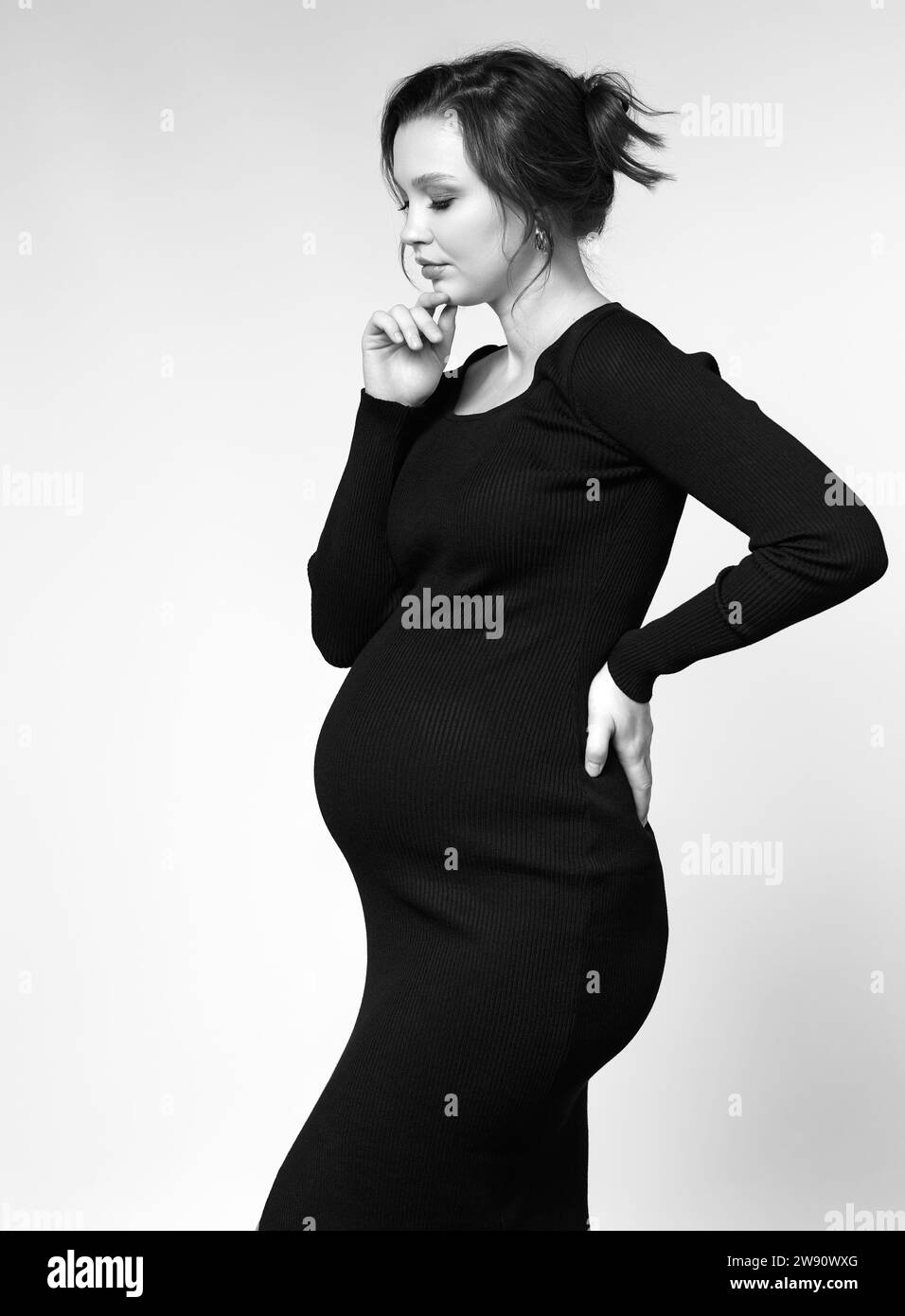 Black and white portrait of young pretty pregnant woman on gray studio background. Female in black dress. Stock Photo