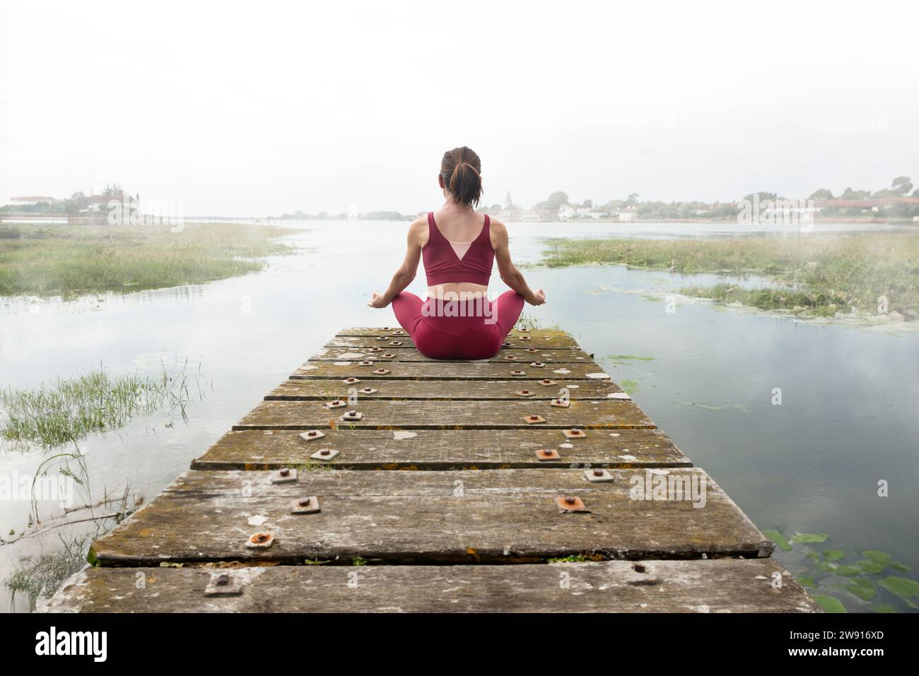 Woman meditating on jetty by lake or river, rear view, mist and fog in the early morning Stock Photo