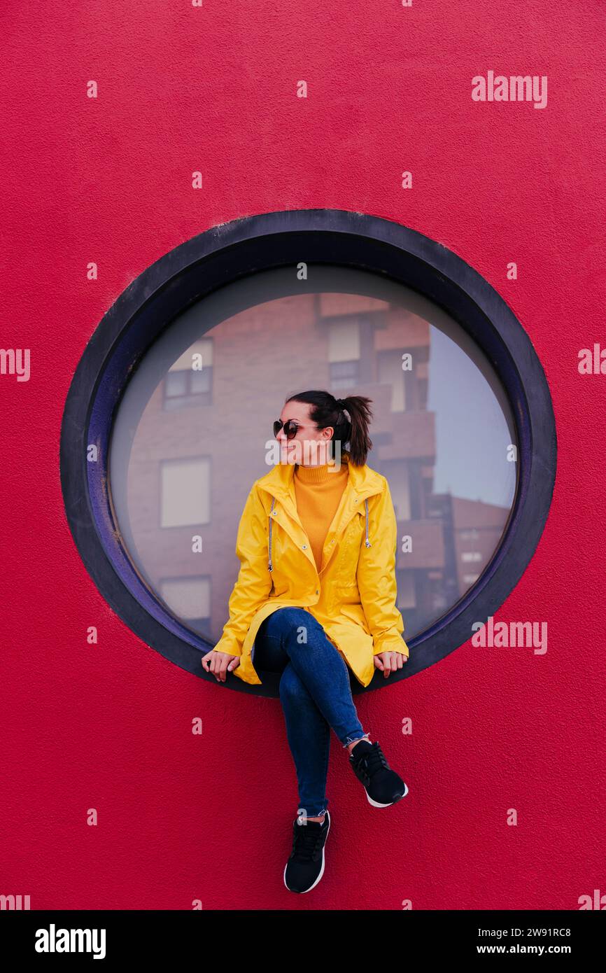 Woman in yellow rain coat sitting in porthole in red wall Stock Photo