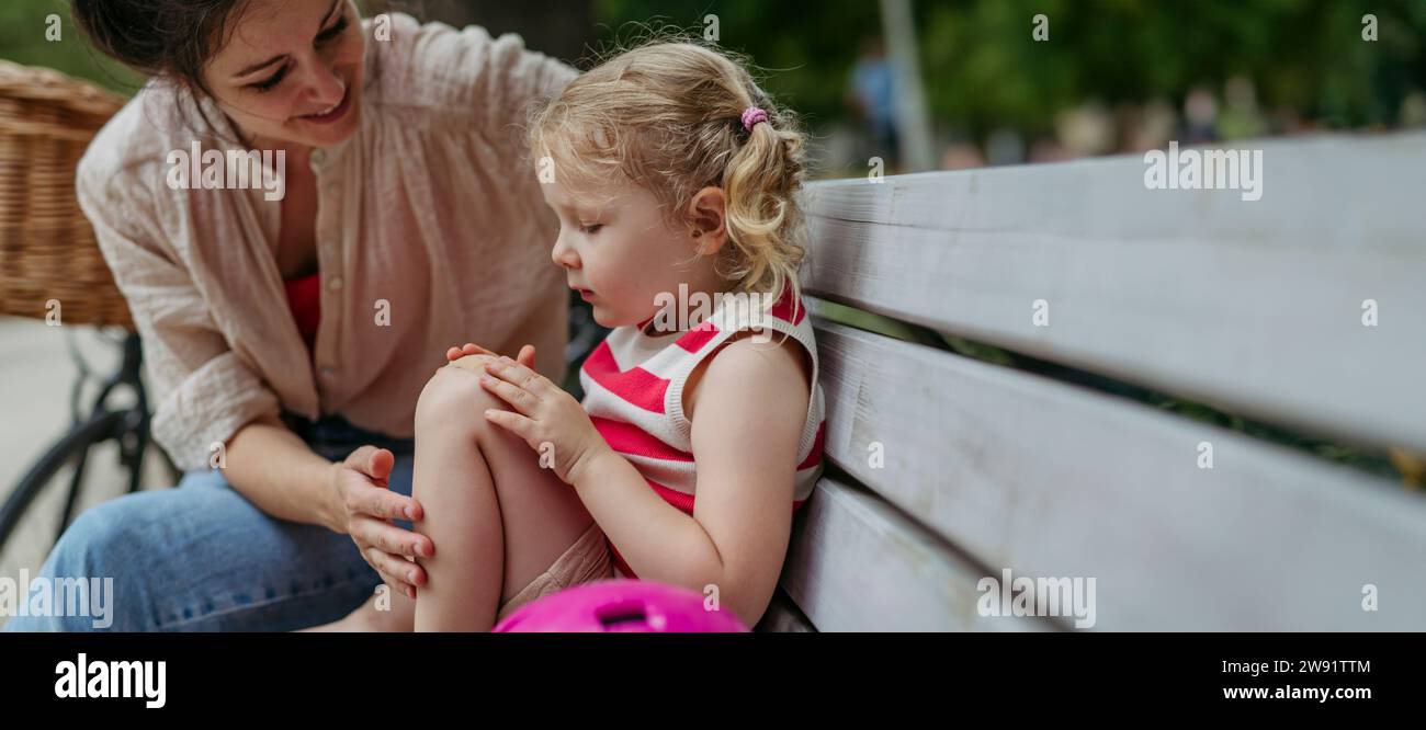 Mother watching little girl blowing on injured knee and putting on band aid Stock Photo