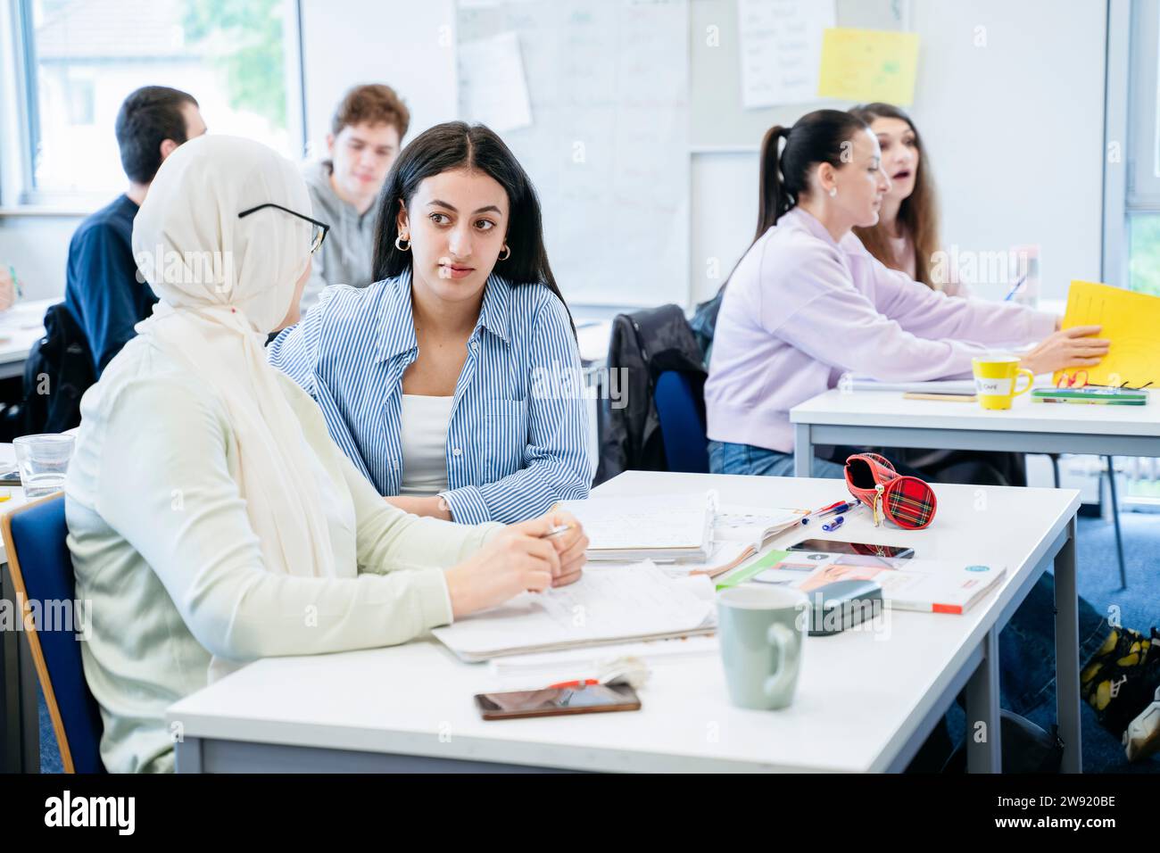 Woman discussing with friend in hijab sitting at desk in classroom Stock Photo