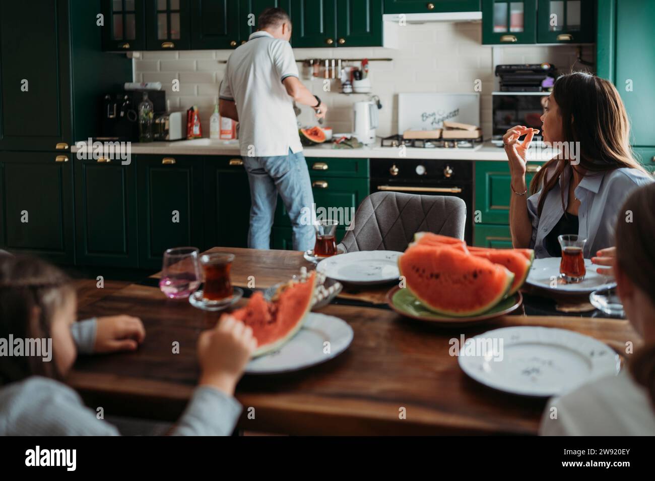 Mother and daughters having lunch at table with man cutting watermelon in kitchen Stock Photo