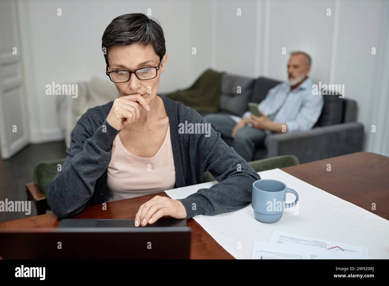 Woman using laptop working from home husband sitting on sofa in background Stock Photo