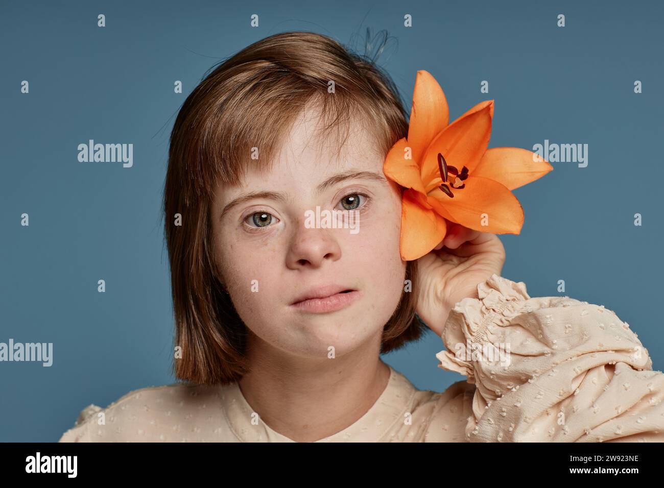 Girl with down syndrome holding orange orchid behind ear against blue background Stock Photo