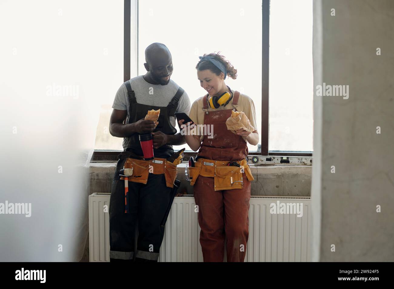 Smiling construction workers taking lunch break and using smart phone at site Stock Photo