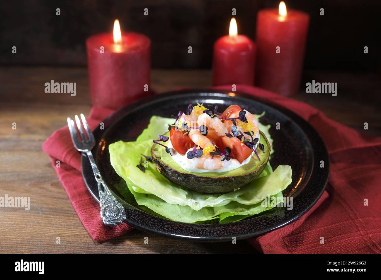 Halved avocado filled with shrimps, tomatoes and mayonnaise on a black plate, red candles and napkin, dark rustic wooden background, festive holiday a Stock Photo
