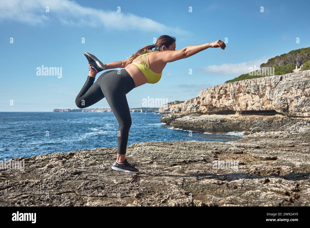 A woman performing a yoga pose on a rocky shore by the sea under a clear sky Stock Photo
