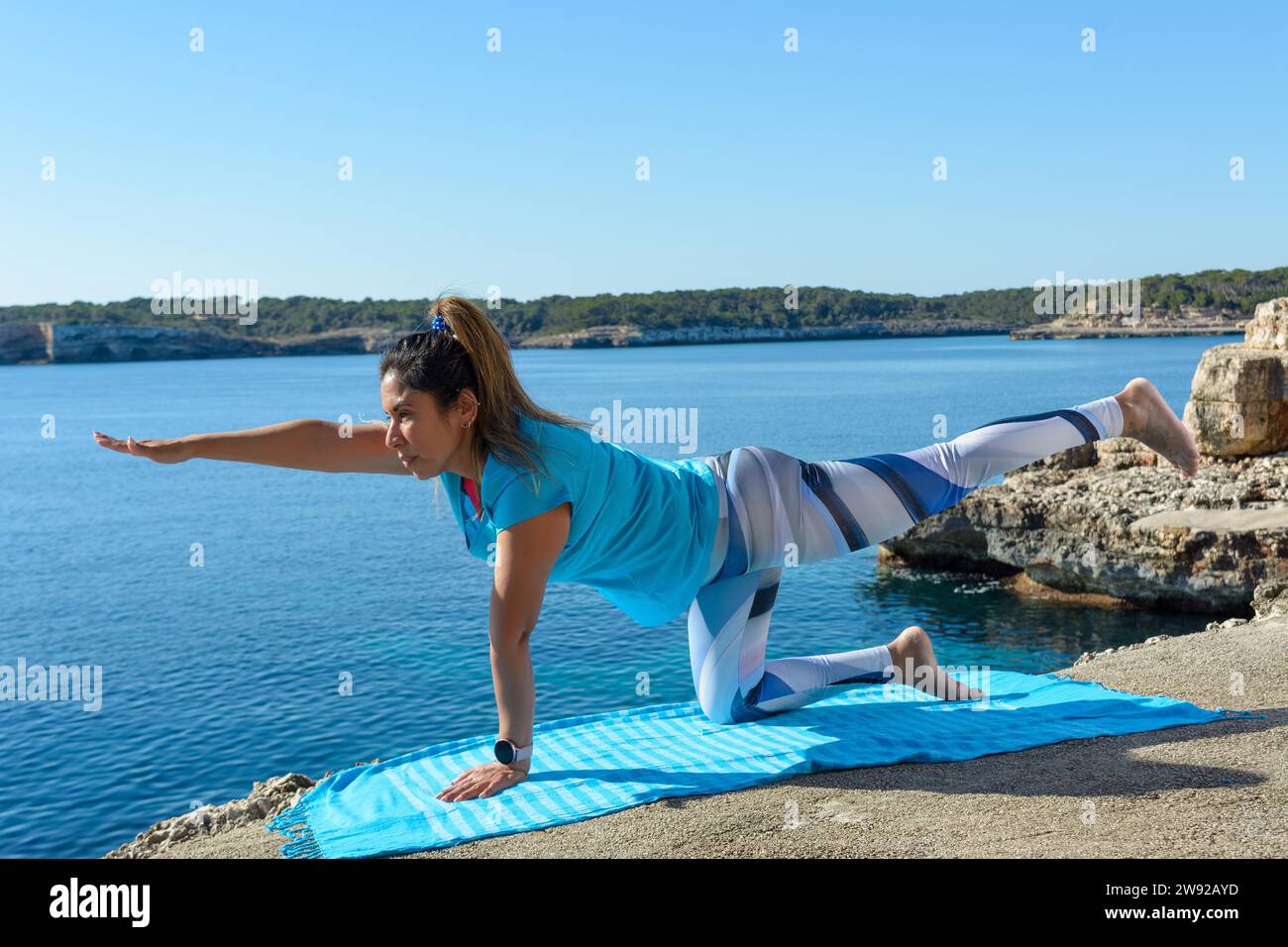A woman performing a yoga balance pose on a mat by the sea on a sunny day, with cliffs under a blue sky Stock Photo