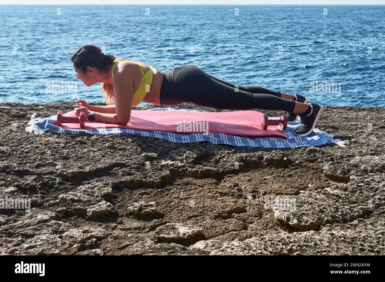 A woman is performing a yoga pose on a mat by the sea on a sunny day Stock Photo