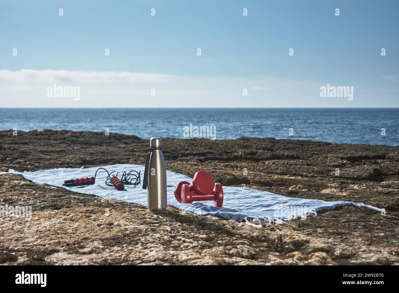 Fitness and sports concept background. Getting ready. Towel, jump rope, dumbbell and bottle of water, by the sea on a sunny winter day, with Stock Photo