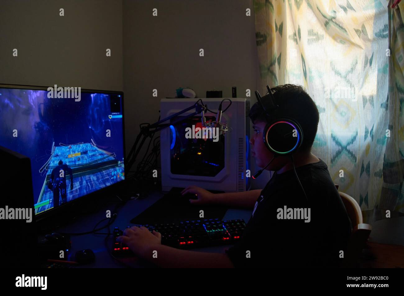 Side View of teenage gamer boy playing video games online on computer in dark room, wearing headphones with microphone and using backlit colorful Stock Photo