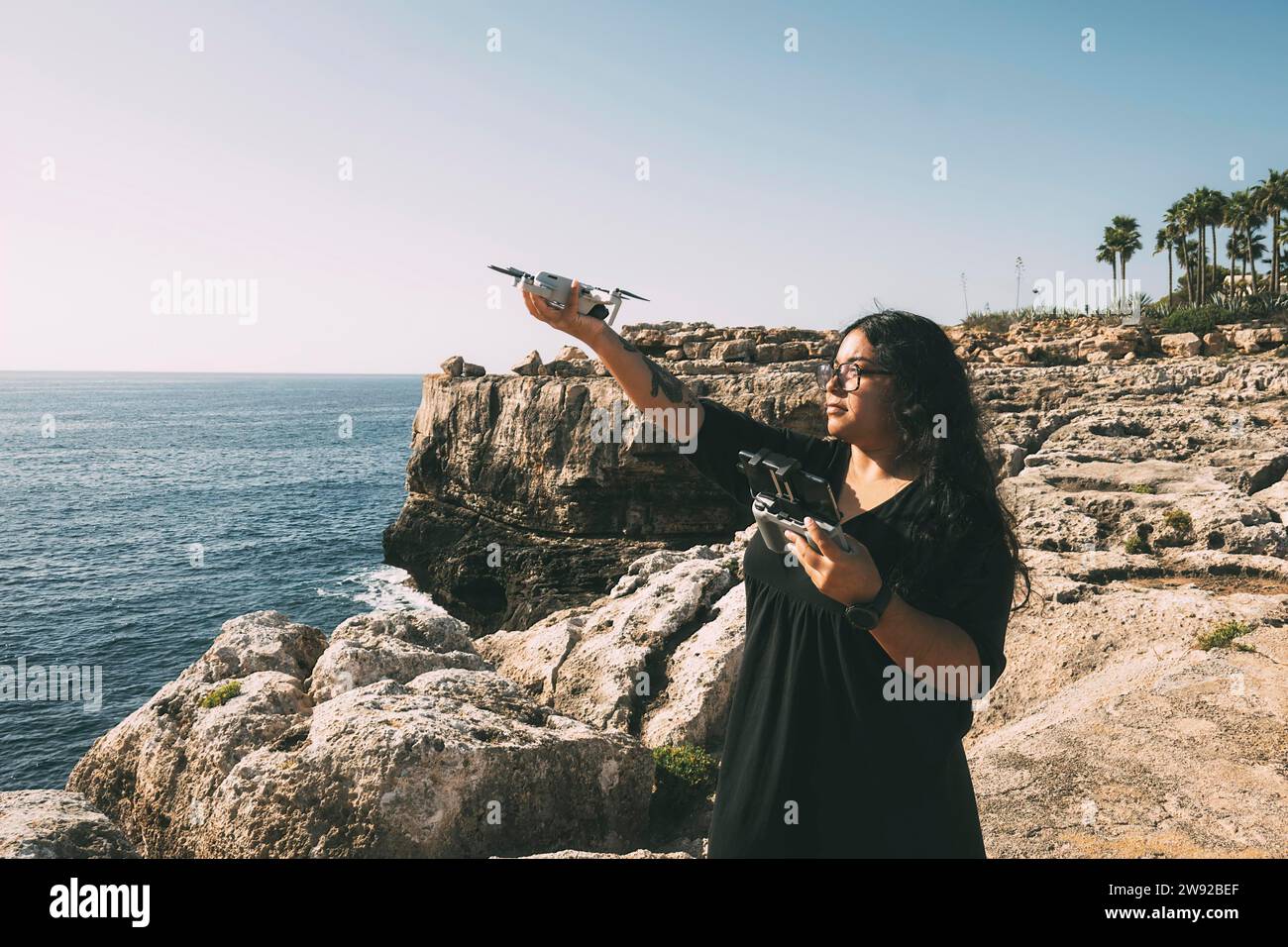 Happy woman with drone, looking to the side black dress and remote control, on cliff, by the sea sunny day. technological concept Stock Photo