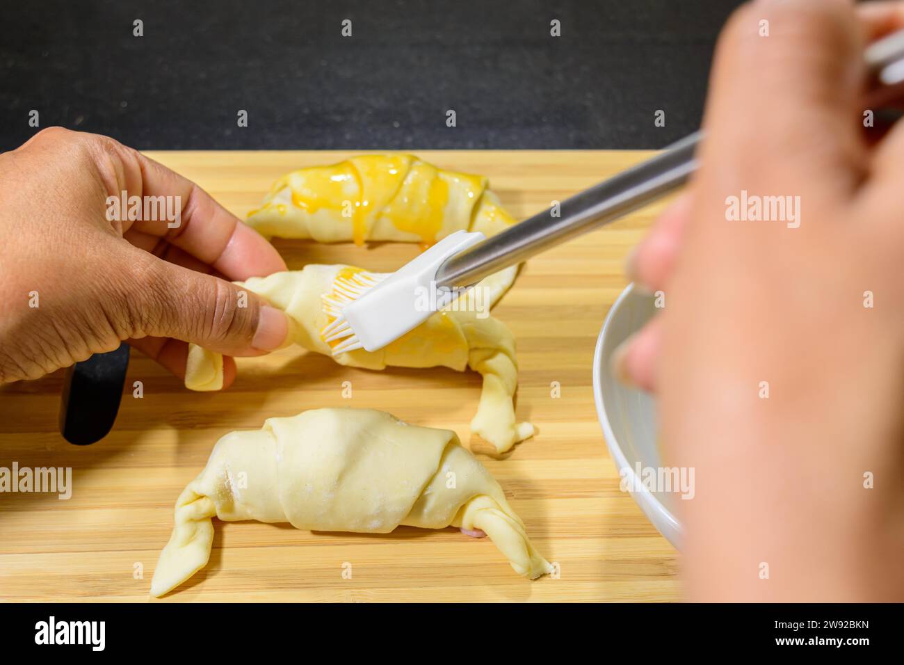 Preparation of croissants, stuffed with ham and cheese and painted with egg yolk, on a wooden board, ready to bake Stock Photo