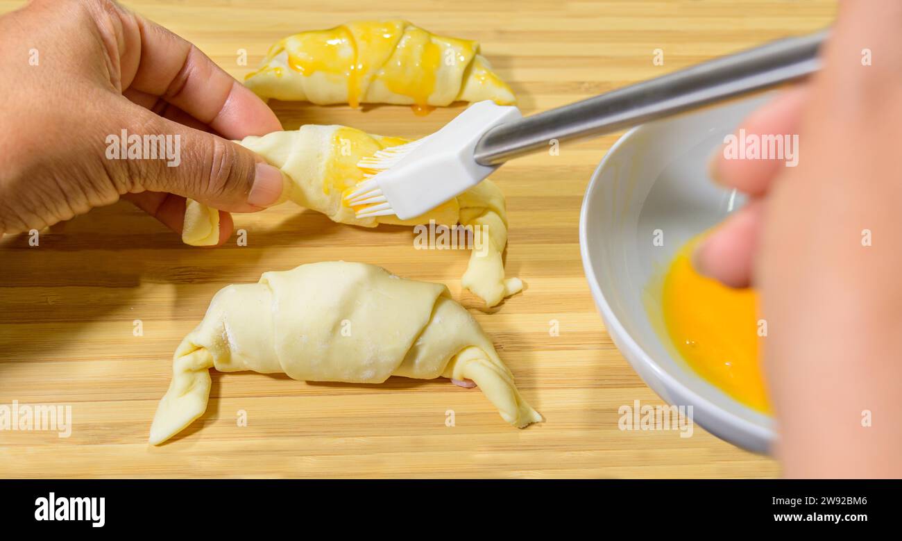 Preparation of croissants, stuffed with ham and cheese and painted with egg yolk, on a wooden board, ready to bake Stock Photo