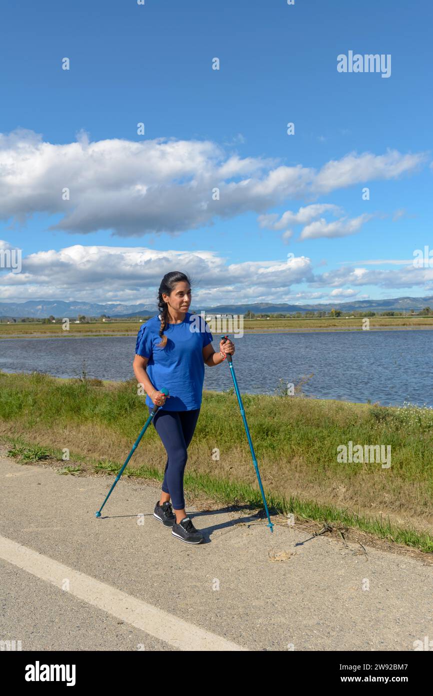 Outdoor enthusiast with walking poles beside a tranquil water body on a bright day, Hispanic Latina woman walking with trekking poles in the Ebro Stock Photo