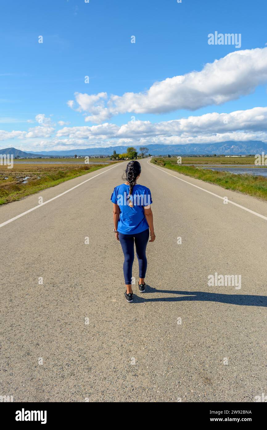 View from behind an individual walking along a deserted road with scenic views on a clear day, back view of latina woman dressed in blue walking down Stock Photo