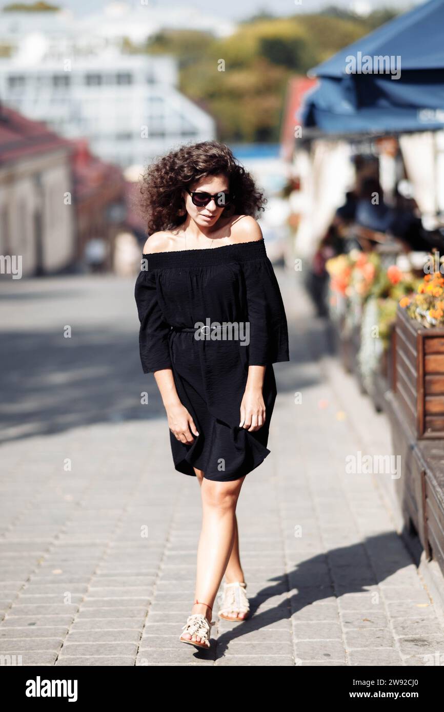 Stylish slender curly woman in a little black dress and sunglasses walks down the street on a sunny day Stock Photo