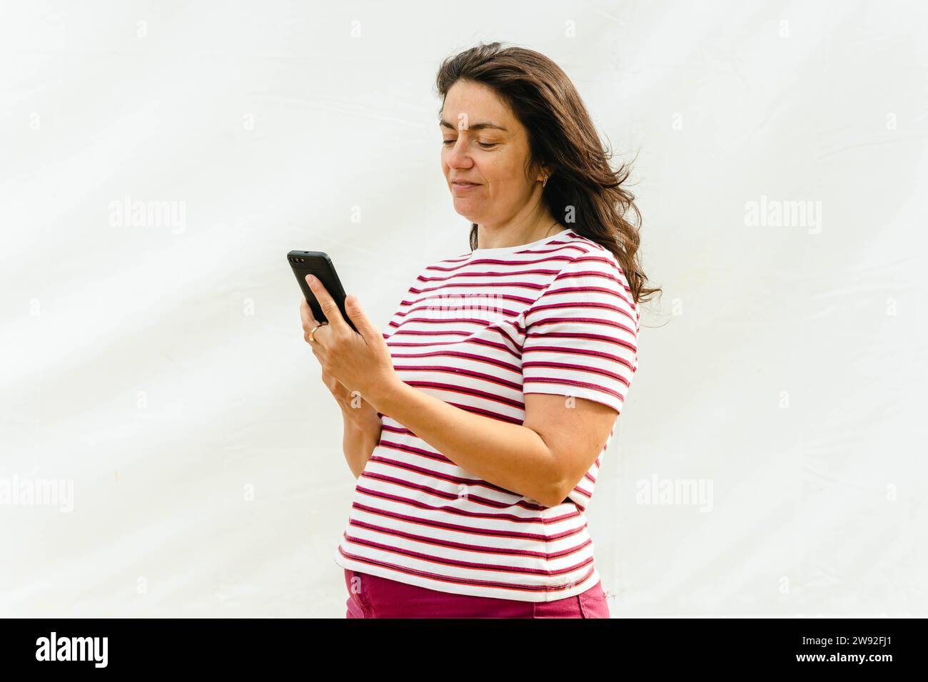 long-haired middle-aged lady in a striped top, engrossed in using her smartphone, exuding a modern and tech-savvy vibe. Stock Photo