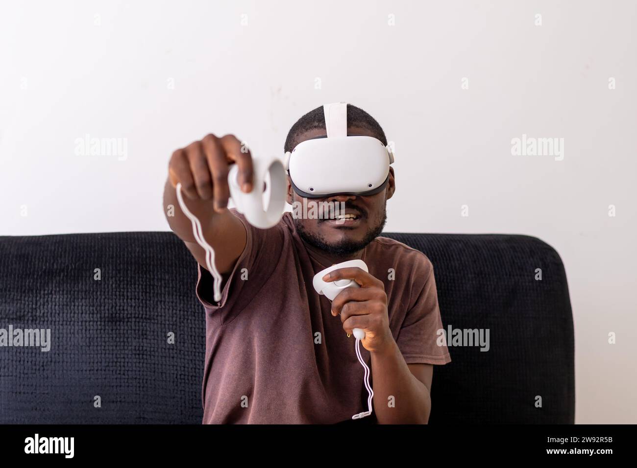 young man wearing a vr headset engrossed in a virtual reality game and holding controllers sitting on a couch. Vr gaming concept Stock Photo