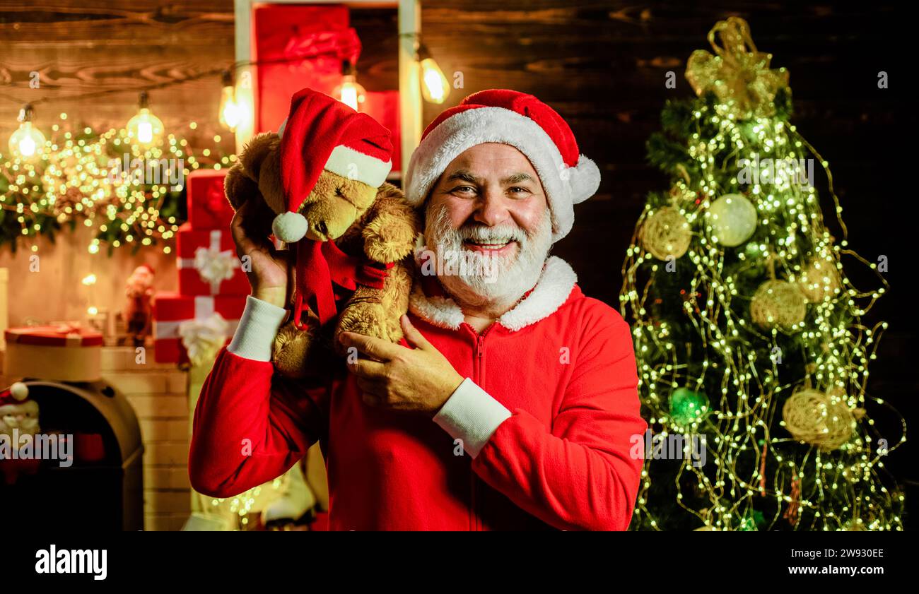 Smiling Santa Claus with plush teddy bear on shoulder in room decorated for Christmas. New Year holidays. Christmas background. Bearded man in Santa Stock Photo