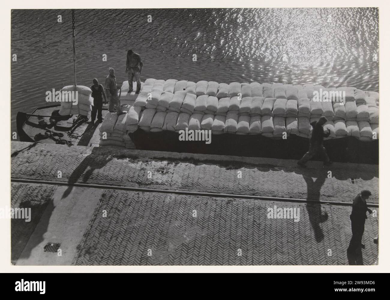 Allied food aid: the unloading of a deck barge, 1945 documentary photographs On a quay in the Rotterdam (?) Haven, a full deck barge is loaded with bags unloaded by three port workers. The first pockets are hoisted from the deck barge against the background of the water and with the quay with ankle track in the foreground. The deck barges sailed from the Allied 'Liberty' cargo ships to the warehouses where the food from England and America was stored.  photographic support Stock Photo