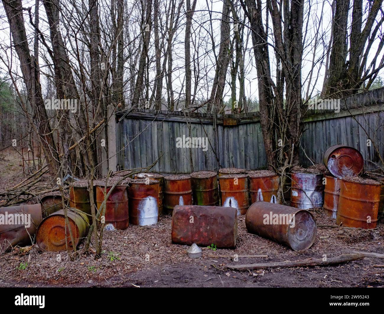 This image shows rusty barrels that are scattered in front of a wooden fence. Stock Photo
