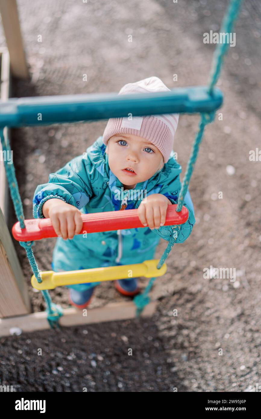 Little girl holds on to the rungs of a rope ladder and looks up. Top view Stock Photo