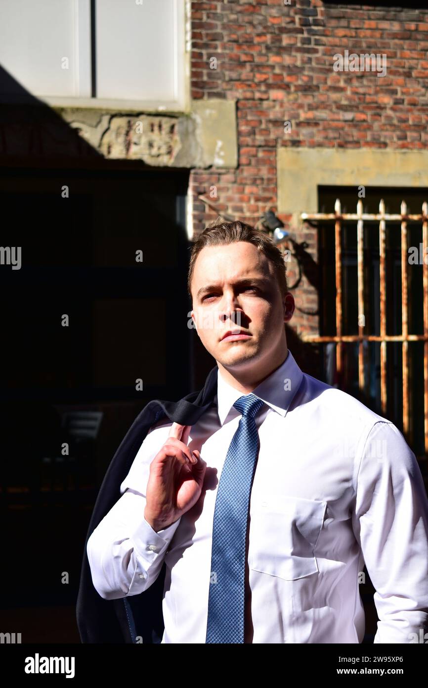 Young muscular white man carrying his black suit jacket over his shoulder and wearing a white collared shirt and blue tie in front of a brick wall Stock Photo