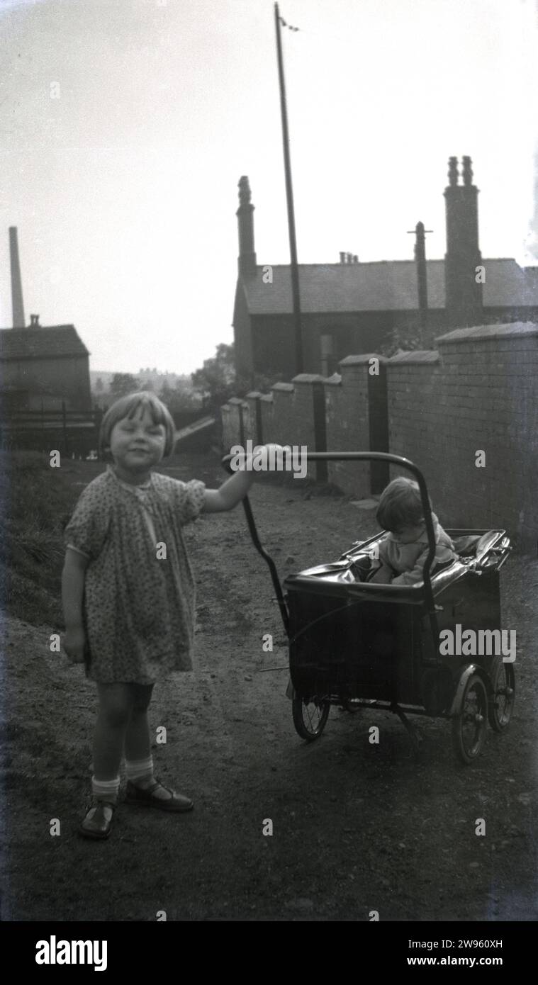 1950s, historical, post-war Manchester, a scruffy little girl holding the handle of a small pram with her infant brother on a gravel alley at the back of a row of victorian terraced houses, England, UK. Stock Photo