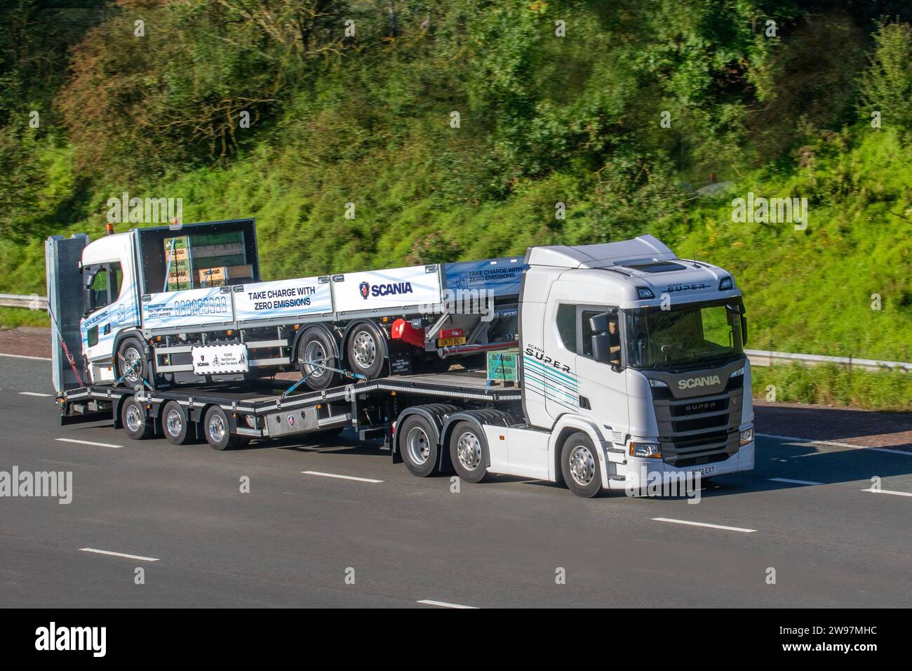 100% Scania L Series (6) Electric Collier flatbed truck body with Anteo Tail Lift carried piggy-back on 2023 Scania Super R500 A 6x2/2 Diesel powertrain; travelling at speed on the M6 motorway in Greater Manchester, UK Stock Photo