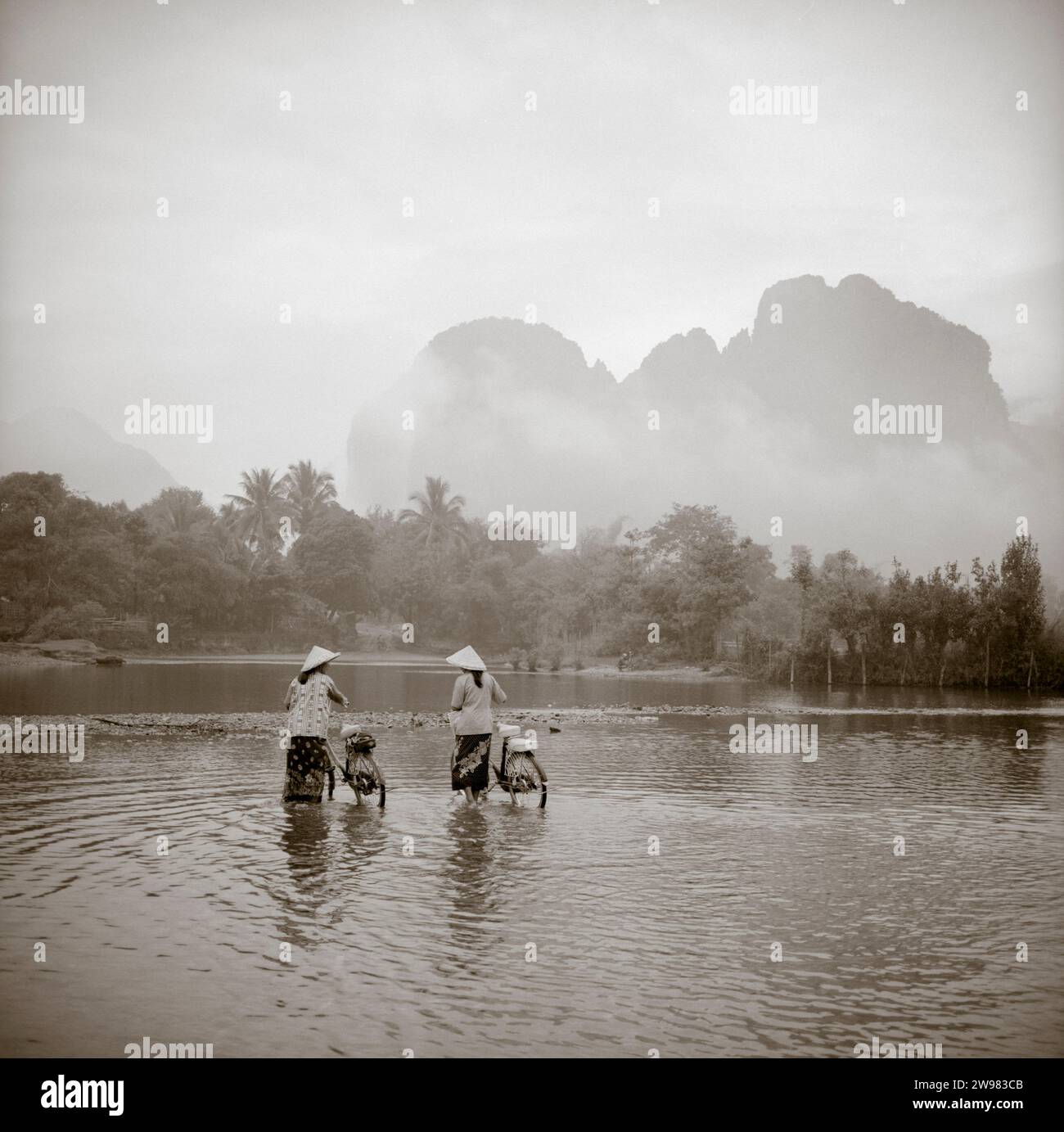 Two Women Wearing Conical Asian Hats Push Bicycles Through A Shallow