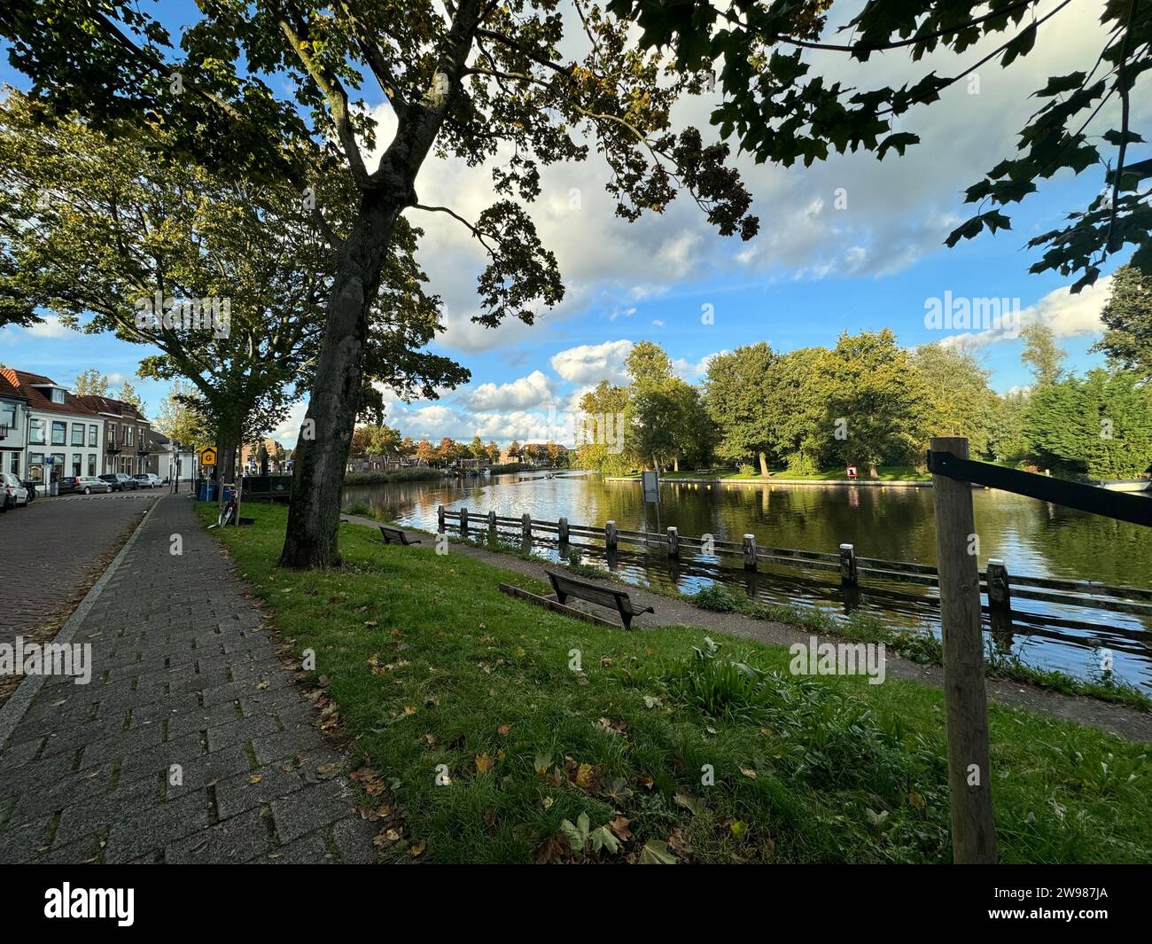 A wooden bench is situated alongside a road that overlooks a tranquil river Stock Photo