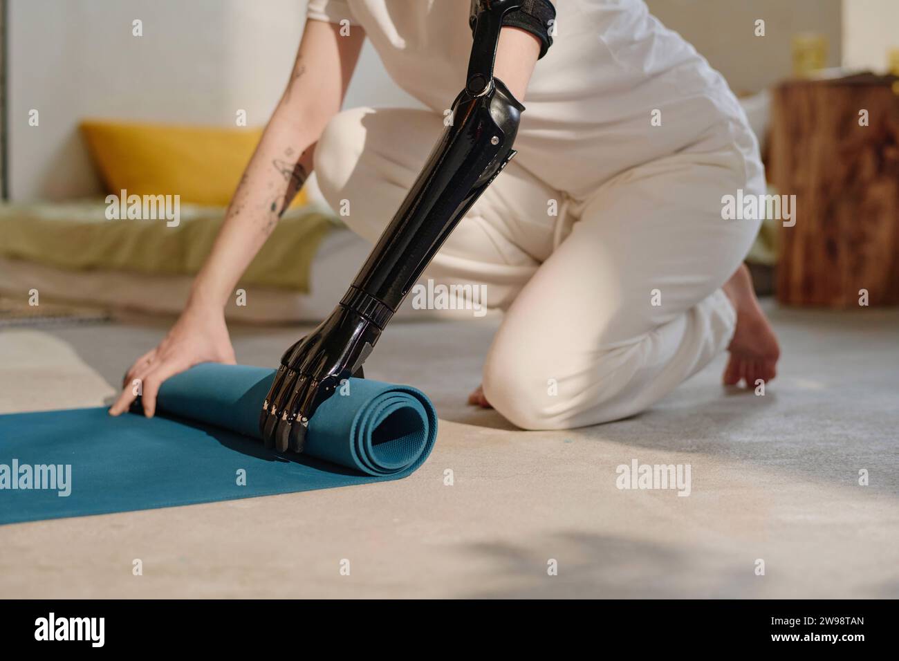Young woman with prosthetic arm rolling out yoga mat to exercise at home Stock Photo