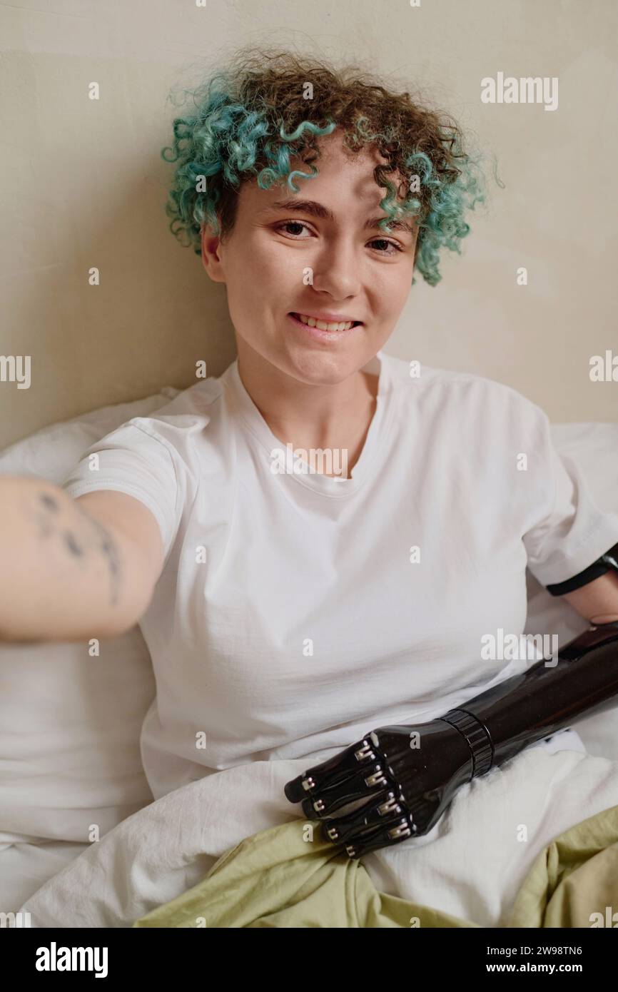 Smiling curly young woman with prosthetic arm taking selfie in bed Stock Photo