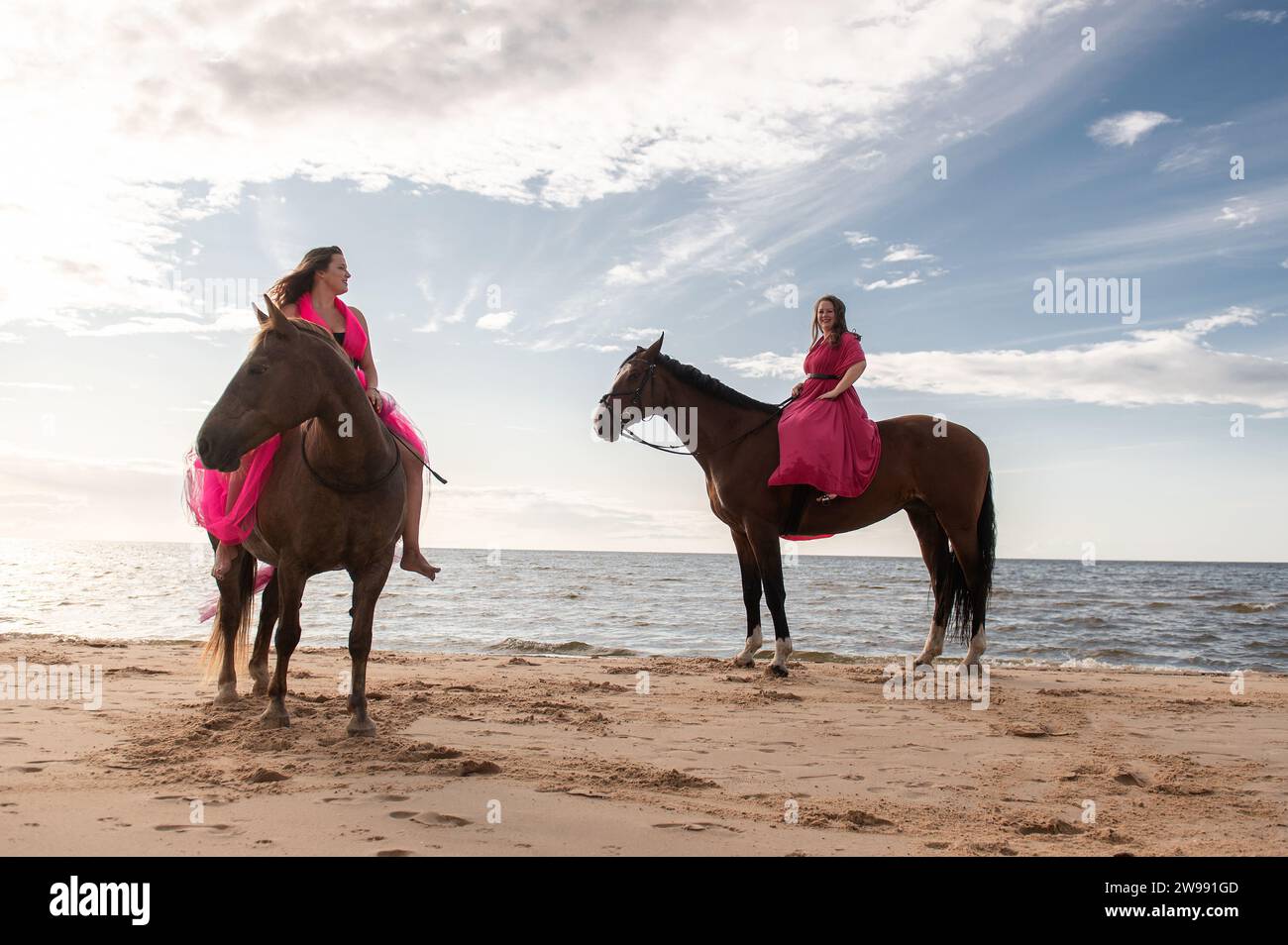 A pair of young female friends in matching dresses posing on horses standing on a beach Stock Photo