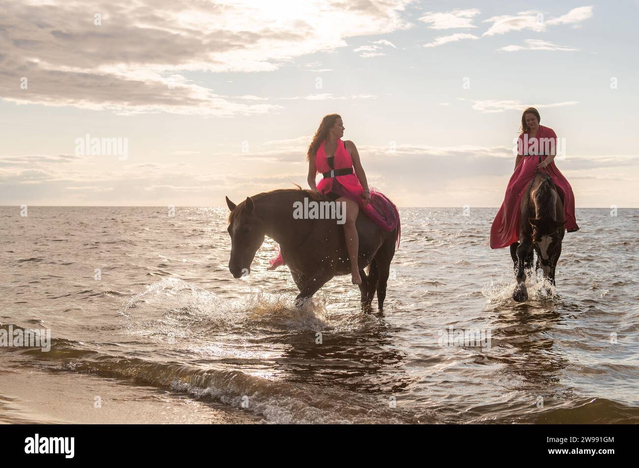 A pair of young female friends in matching dresses posing on horses standing on a beach Stock Photo