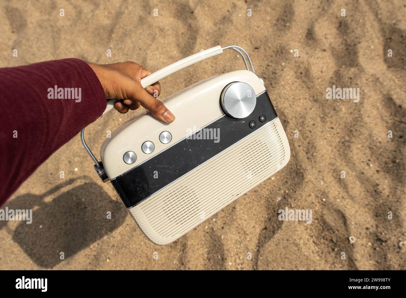 A young adult stands on a sandy beach, gesturing towards an old-fashioned radio sitting nearby Stock Photo